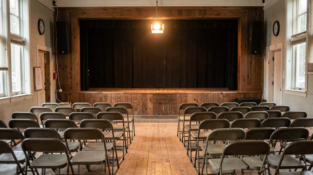 A small-town community theatre stage lit by a single warm work light, rows of folding chairs in the foreground, framed straight-on from the empty audience seats.